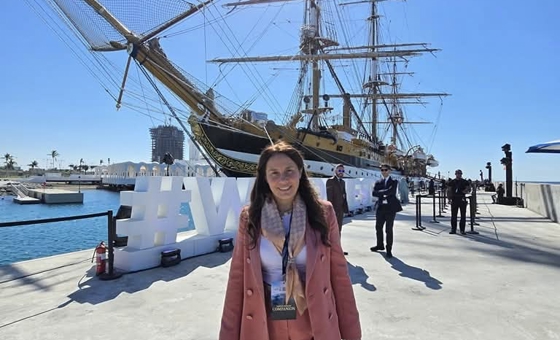 Alessandra Locatelli sul molo davanti alla nave scuola Amerigo Vespucci ormeggiata in porto; sullo sfondo sono visibili gli alberi dell'imbarcazione e le strutture portuali.
